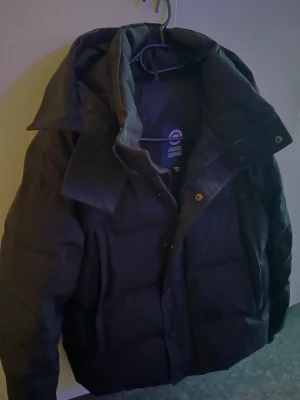 Svart dunjacka - Säljer en svart dunjacka med hög krage och huva. Jackan har dragkedja och knappar framtill, som håller värmen. Perfekt för alla väder.