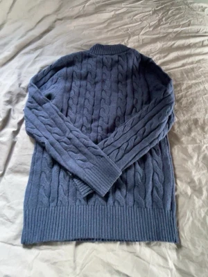 Mörkblå kabelstickad tröja Polo Ralph Lauren - Mörkblå kabelstickad tröja från Polo Ralph Lauren med halv dragkedja och röd broderad logga på bröstet. Tröjan har ribbade muddar vid ärmslut och nederkant samt hög krage. Perfekt för kyliga dagar och snygg till jeans.