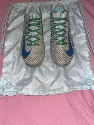 Nike Mercurial fotbollsskor elite ljusblåa - Säljer ett par Nike Mercurial fotbollsskor i silver med blå detaljer och gröna skosnören. Skorna har en modern, strömlinjeformad design och är tillverkade i syntetmaterial för bästa grepp och känsla på planen. Sulan har blå dobbar för bra fäste på gräs.