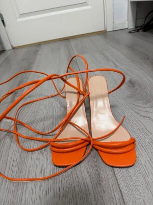 Orange pumps med snörning - Säljer ett par trendiga orange pumps med långa snören att knyta runt ankeln. Skorna har fyrkantig tå och tunna remmar framtill. Klacken är smal och ger en elegant look. Perfekta för dig som vill sticka ut med färg och stil.