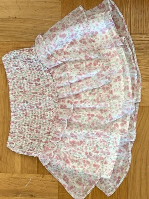Blommig volangkjol från Gina Tricot - Supersöt kort kjol från Gina Tricot med blommigt mönster i rosa, grönt och vitt. Kjolen har volanger och smock i midjan för en bekväm och snygg passform. 