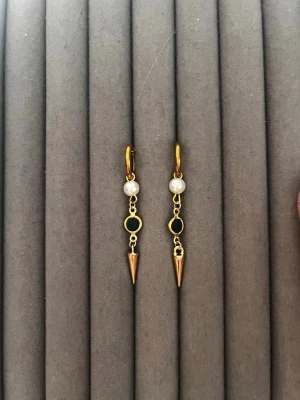 Guldörhängen med pärlor och svarta detaljer - Säljer ett par trendiga örhängen i guldfärg med vita pärlor, svarta runda detaljer och spetsiga hängen. Örhängena har en modern design med mix av material och färger som ger en cool kontrast. Perfekta för dig som gillar unika accessoarer.