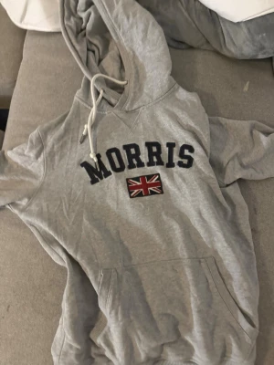 Grå hoodie från Morris - Snygg grå hoodie från Morris med stor huva och dragsko. Framtill står det 'MORRIS' i svart text och under finns en brittisk flagga. Hoodien har en klassisk känguruficka och är gjord i mjukt bomullsmaterial.