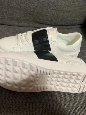 Valentino Garavani vita sneakers 42 - Snygga vita sneakers från Valentino Garavani med en bred svart rem över mitten och ikoniska nitar på hälen. Skorna är i slätt läder och har vita snören samt en chunky sula med rutmönster undertill. Perfekta för dig som gillar exklusiv streetstyle.