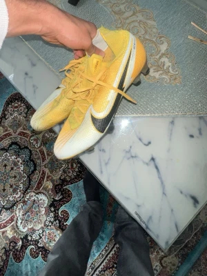 Nike Mercurial gula fotbollsskor - Säljer ett par Nike Mercurial fotbollsskor i gult och vitt med svarta detaljer. Skorna har snörning, låg profil och slitstarka dobbar för bra grepp på planen. Tillverkade i syntetmaterial för lätt känsla och optimal bollkontroll. Perfekta för snabba spelare som vill sticka ut.