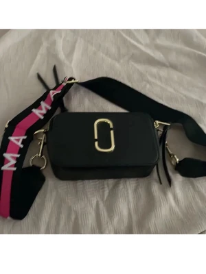Svart axelväska Marc Jacobs - Snygg svart axelväska från Marc Jacobs med guldiga metalldetaljer och bred axelrem i svart och rosa med vit text. Väskan har en rektangulär form och dragkedja upptill. Perfekt för dig som vill ha en trendig och praktisk väska.