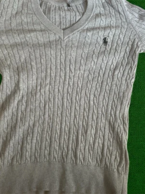 Ljusgrå kabelstickad tröja Polo Ralph Lauren - Kabelstickad tröja i ljusgrått från Polo Ralph Lauren med V-ringning och klassisk broderad logga på bröstet. Tröjan har långa ärmar och ribbade muddar vid ärmslut och nederkant. Perfekt för lager på lager och stilrena outfits. 