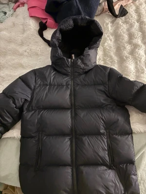 Svart pufferjacka med huva från colmar - Säljer en svart pufferjacka med huva och dragkedja framtill. Jackan har två sidofickor med dragkedja och breda, vadderade paneler för extra värme. Perfekt för kalla dagar och riktigt snygg till streetwear-stilen.