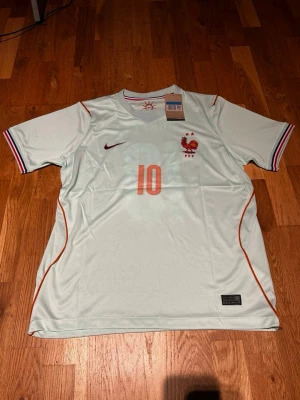 France WC 26 away kit Mbappe #10 - Ljusblå fotbollströja från Nike med orange detaljer, nummer 10 och Mbappé på ryggen. Tröjan har korta ärmar, V-ringning och tunna ränder i orange längs sidorna. Franskt landslagsmärke på bröstet och randiga muddar vid ärmsluten.