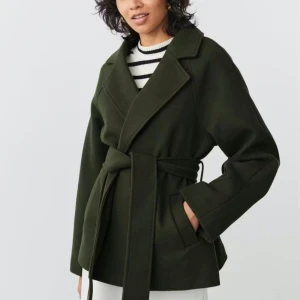 Mörkgrön kappa med knytskärp - Stilren mörkgrön kappa med bred krage och knytskärp i midjan. Kappan har raka ärmar och två sidofickor. Perfekt för dig som gillar en minimalistisk look och vill ha en snygg siluett. Materialet ser ut att vara ullblandning.