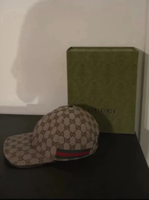 Gucci beige GG keps med röd-grön rand - Säljer en beige keps från Gucci med det klassiska GG-monogrammet över hela kepsen. På sidan finns en ikonisk röd och grön rand. Kepsen har böjd skärm och justerbar rem bak. Materialet är canvas med insida i tyg. Perfekt för dig som vill ha en lyxig och trendig accessoar.