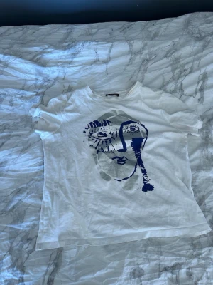 Vit t-shirt med konstnärligt ansikte - Vit t-shirt med ett stort, abstrakt ansikte tryckt i blått och grått på framsidan. T-shirten har korta ärmar och en klassisk rund halsringning. Perfekt för dig som gillar unika och kreativa prints.