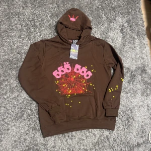 Brun hoodie med rosa 555-tryck - Cool brun hoodie från 555 Soul med stor rosa 555-text på bröstet, röd spindelnätsgrafik och gula stjärnor. Huva med tryck och känguruficka framtill. Perfekt för dig som gillar streetwear och unika prints. Materialet känns mjukt och skönt, troligen bomull.