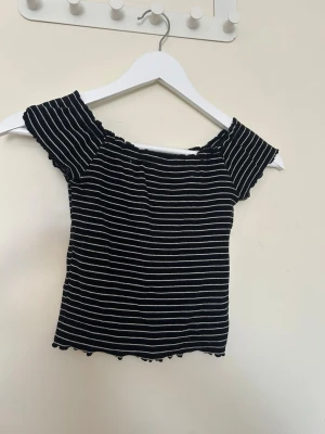 Svart ribbad offshoulder topp Gina Tricot - Säljer en svart ribbad offshoulder topp från Gina Tricot i storlek XS. Toppen har vita tunna ränder, stretchigt material och vågig kant nertill och vid ärmarna. Perfekt för dig som gillar en croppad och figurnära stil.