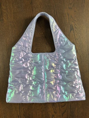 Hvisk Quilted Handbag Dam - Väska från Hvisk i quiltad, iriserande material med skimrande nyanser av lila, grönt och rosa. Väskan har en bred, rektangulär form och ett stort handtag.