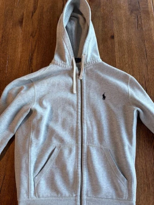 Grå hoodie från Polo Ralph Lauren - Snygg grå hoodie från Polo Ralph Lauren med dragkedja framtill och två fickor. Klassisk broderad logga på bröstet och justerbar huva med snören. Tillverkad i mjukt bomullsmaterial som är perfekt för chill dagar.