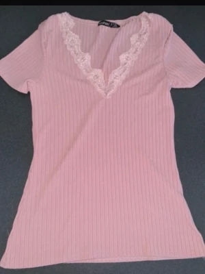 Topp - Söt ljusrosa ribbad topp med korta ärmar och djup V-ringning dekorerad med vit spets. Materialet är stretchigt och mjukt, vilket ger en bekväm passform. Perfekt för en trendig och feminin look.