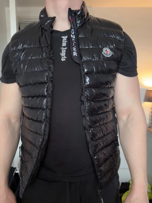 Svart dunväst från Moncler - Snygg svart dunväst från Moncler med glansig finish och klassisk logga på bröstet. Västen har hög krage, dragkedja framtill och diskreta sidofickor. 