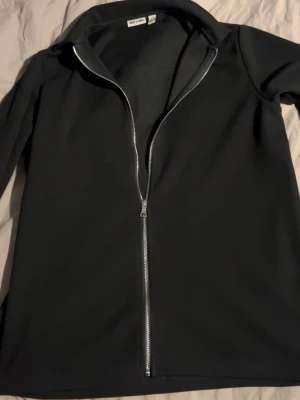 Svart overshirt från Only & Sons - Stilren svart overshirt från Only & Sons med hel dragkedja framtill och klassisk krage. Materialet är mjukt och har en slät finish, perfekt för lager-på-lager. Enkel design utan fickor eller mönster, passar dig som gillar minimalistiskt. Aldrig använd!