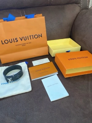 Louis Vuitton svart monogram armband - Snyggt svart armband från Louis Vuitton med klassiskt monogrammönster och silverfärgat LV-spänne. Armbandet är gjort i läder och har en modern, stilren design som passar perfekt till din outfit. Kommer med originalförpackning och dustbag.