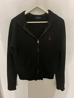 Svart hoodie från Polo Ralph Lauren - Svart hoodie med dragkedja från Polo Ralph Lauren. Klassisk design med huva, kängurufickor och en röd logga på bröstet. Tycker inte alls storleken stämmer. Är normalt S/M 