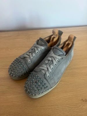 Gråa Loubs sneakers med nitar i mocka - Unika grå sneakers från Christian Loubitouin mocka med coola nitar på tån. En har låstnat. matchande grå skosnören. Skorna har en låg profil och en klassisk sula i vitt. Perfekta för dig som vill sticka ut med en edgy och trendig stil.
