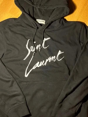 Svart Saint Laurent hoodie med tryck - Svart hoodie från Saint Laurent med vit logotyp i handskriven stil på bröstet. Skitsnygg o perfekt nu t våren🤩. Strl M. Hör av dig om du har några frågor!💯
