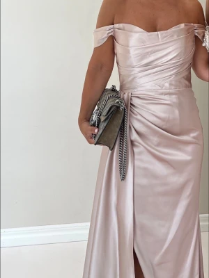 Beige axelbandslös långklänning  - Elegant beige långklänning i satinliknande material med offshoulder och draperad överdel. Klänningen har en slits framtill och smickrande omlottdetalj i midjan. Perfekt för speciella tillfällen.