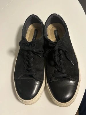 Svarta sneakers från Sneaky Steve - Säljer ett par svarta sneakers från Sneaky Steve med mockaliknande ovandel och blanka tåhättor i svart. Skorna har vita sulor och klassisk snörning. Perfekta för dig som gillar stilrena och tidlösa sneakers med en twist.