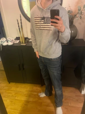 Grå hoodie med amerikansk flagga - Säljer en grå hoodie med huva och dragsnören. På bröstet finns ett tryck av den amerikanska flaggan i en lite sliten stil. Hoodien har en klassisk känguruficka framtill och är gjord i mjukt sweatshirtmaterial. Perfekt för en avslappnad och cool look.