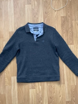 Mörkgrå tröja med krage - Mörkgrå långärmad tröja från med klassisk krage och knappslå. Tröjan har en diskret logga på bröstet och är tillverkad i mjukt material som passar perfekt till en avslappnad stil.