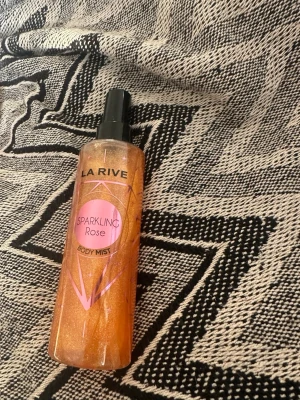 La Rive Sparkling sparkling Rose Body Mist orange glittrig - Fräsch body mist från La Rive med doft av Sparkling Rose. Flaskan är genomskinlig med skimrande rosa-orange vätska och svart spraykork. Perfekt för dig som gillar blommiga och fruktiga dofter med en touch av glitter. Body listen är knappt använd max 2 ggr 💗 200 ml