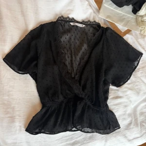 Svart spetts blus från Zara - Snygg svart transparent blus från Zara med omlott-design och spetsdetaljer vid halsringningen. Blusen har korta ärmar och ett diskret prickigt mönster i tyget. Resår i midjan ger en feminin siluett. Perfekt att styla lager på lager.