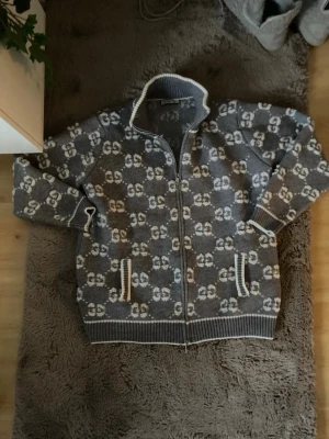 Grå ziptröja med GG-mönster från G0cci - Säljer en grå stickad ziptröja från Gg med ikoniskt GG-mönster i vitt över hela tröjan. Tröjan har hög krage, dragkedja framtill och två fickor med vita detaljer. Perfekt för dig som vill ha en lyxig och trendig look. Passsar L XL