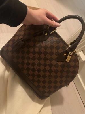 Louis Vuitton Damier Alma PM Väska - Säljer en ikonisk handväska från Louis Vuitton med klassiskt brunt och mörkbrunt rutigt mönster. Har några repor samt fläck i insidan men inte något som märks så mycket. Vid intresse kan jag skicka fler bilder videos.