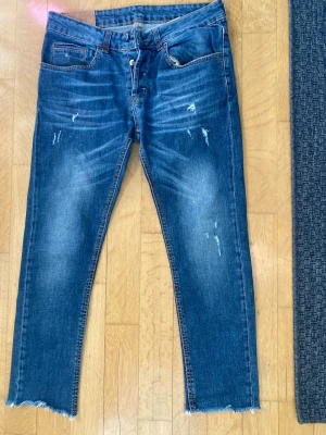 Blå slitna jeans från Dondup - Säljer ett par blå jeans från Dondup med slitningar och råa kanter nertill. Jeansen har klassisk femficksmodell, normal midja och är tillverkade i denim. Snygg tvätt med ljusare partier och trendiga detaljer som ger en cool look.