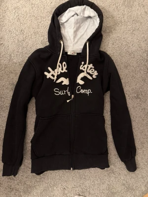 Svart Hollister hoodie med dragkedja - Svart hoodie från Hollister med vit broderad text och logga på bröstet. Dragkedja framtill, två fickor och stora vita snören i huvan. Insidan av huvan är ljusgrå och materialet är mjukt och skönt, perfekt för chill dagar.
