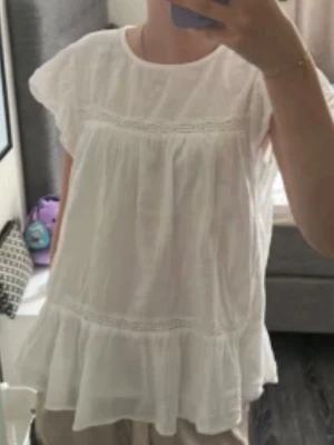 Vit volangblus Gina Tricot  - Söt vit blus med volangkant nedtill och fina spetsdetaljer över bröstet och vid midjan. Blusen har en lös och luftig passform samt korta ärmar. Perfekt för en fräsch och somrig look.