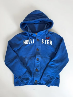 Blå hoodie från Hollister med knappar - Snygg blå hoodie från Hollister med stora vita bokstäver och röd logga på bröstet. Hoodien har knappstängning framtill, två fickor och en rejäl huva. Tillverkad i mjukt bomullsmaterial som känns skönt mot huden. Perfekt för en avslappnad och cool stil.