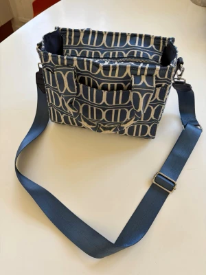 Blå axelväska från Don Donna - Snygg blå axelväska från Don Donna med ikoniskt monogrammönster i vitt. Väskan har justerbar axelrem i textil, flera fack och dragkedja upptill. Perfekt för dig som vill ha en trendig och praktisk väska med lyxig känsla.