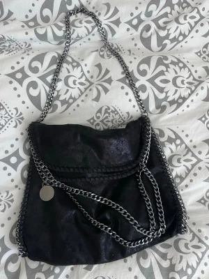 Svart axelväska med kedjedetaljer  - Snygg svart axelväska med silverfärgade kedjor längs kanterna och som axelrem. Väskan har en mjuk, något glansig yta och rundad form. Dekorativ metallbricka framtill. 