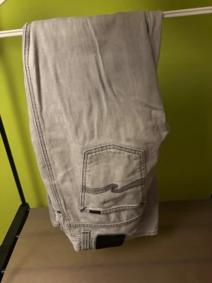 Ljusgrå jeans från Nudie Jeans - Säljer ett par ljusgrå jeans från Nudie Jeans med klassisk femficksdesign och snygg broderad bakficka. Jeansen har raka ben och är tillverkade i mjukt denimtyg. Perfekta för en avslappnad och stilren look.