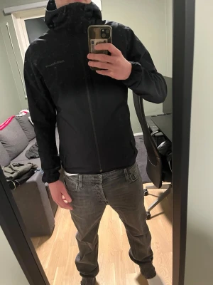 Svart vindjacka från Peak Performance - Snygg svart vindjacka från Peak Performance med huva och dragkedja framtill. Jackan har diskret logga på bröstet och två fickor med dragkedja. Perfekt för blåsiga dagar och enkel att matcha med olika outfits.