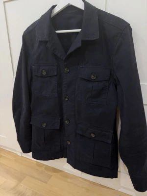 Mörkblå overshirt med fickor - Stilren mörkblå overshirt med fyra stora fickor framtill och knäppning med knappar. Overshirten har klassisk krage och raka ärmar, perfekt för lager-på-lager. Materialet är kraftigt bomullstyg som ger en snygg och avslappnad look.