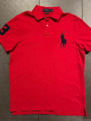 Röd pikétröja från Ralph Lauren - Röd pikétröja från Ralph Lauren med klassisk krage, två knappar och stor marinblå logga på bröstet. Slim fit-modell med siffra 3 på ärmen. Perfekt för en sportig och stilren look.