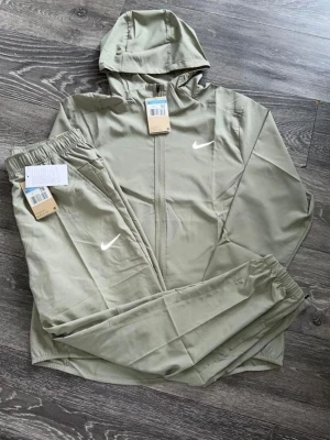 Beige träningsset från Nike - Snyggt beige träningsset från Nike med matchande hoodie och byxor. Setet har dragkedja på tröjan, huva och elastiska muddar på byxorna. Båda delarna har den klassiska vita Nike-loggan. Perfekt för gymmet eller chill.