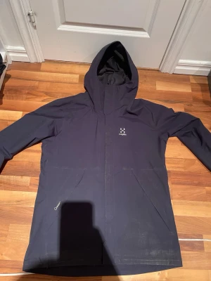 Mörkblå regnjacka från Haglöfs - Mörkblå regnjacka från Haglöfs med huva och diskret logga på bröstet. Jackan har långa ärmar, dragkedja framtill och två snedställda fickor. Tillverkad i slitstark syntet som står emot regn och vind. Perfekt för blöta dagar. Lite smuts men det går bort i tvätten annars perfekt!