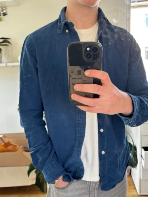Mörkblå skjorta från Ralph Lauren - Säljer en mörkblå skjorta från Ralph Lauren med klassisk button-down krage och broderad logga på bröstet. Skjortan har långa ärmar, vita knappar och är gjord i mjuk bomull. Perfekt att bära öppen över en t-shirt för en avslappnad look.