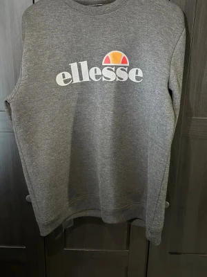 Grå sweatshirt från Ellesse - Snygg grå sweatshirt från Ellesse med klassisk logga i vitt, orange och rött på bröstet. Tröjan har rund halsringning, ribbade muddar vid ärmslut och nederkant samt en avslappnad passform. Perfekt för en casual och sportig stil.