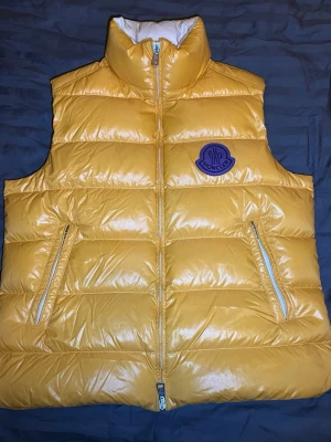 Gul dunväst från Moncler - Snygg gul dunväst från Moncler med hög krage och två dragkedjeförsedda fickor. Västen har ett glansigt yttertyg och ett stort Moncler-märke i lila på bröstet. Perfekt för lager-på-lager och streetwear-vibe. Tveka ej på att höra av dig vid frågor osv.